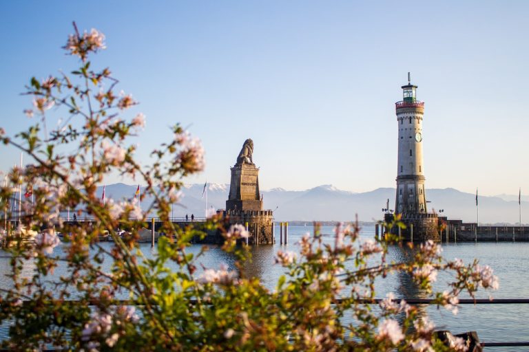 Leuchtturm und bayerischer Löwe am Hafen von Lindau am Bodensee, umrahmt von blühenden Zweigen, mit Blick auf die schneebedeckten Alpen im Hintergrund